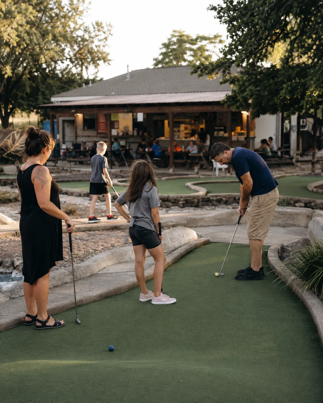 A family mid-round on an outdoor mini golf hole, with a snack counter and patio tables behind them