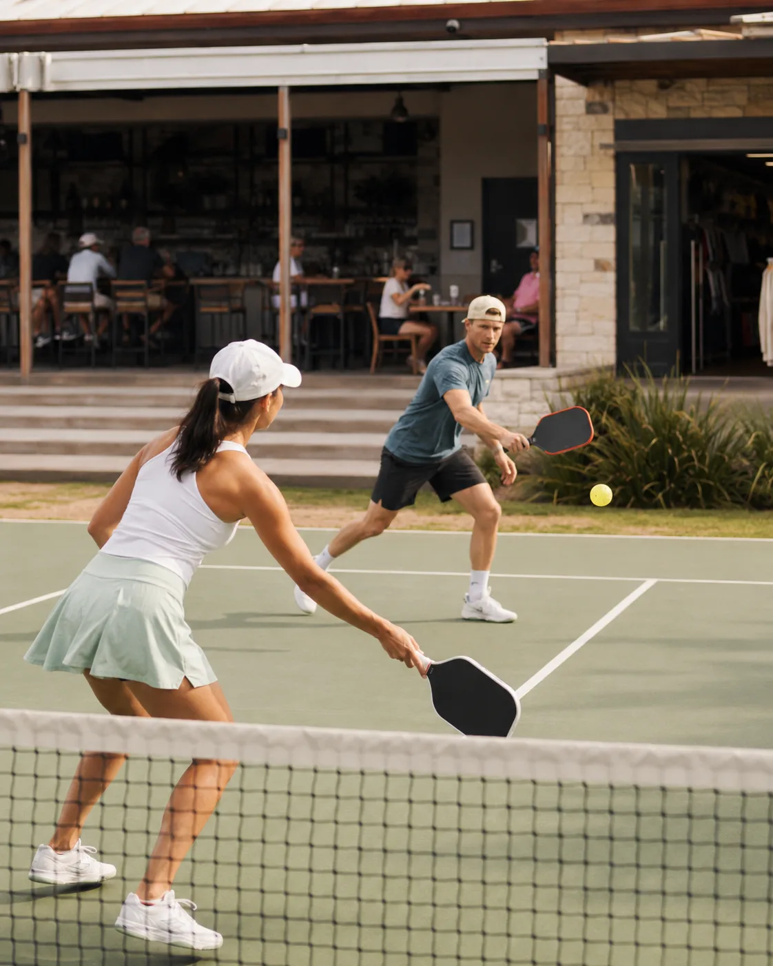 Two players mid-rally on an outdoor pickleball court, with a covered bar patio and pro shop behind them
