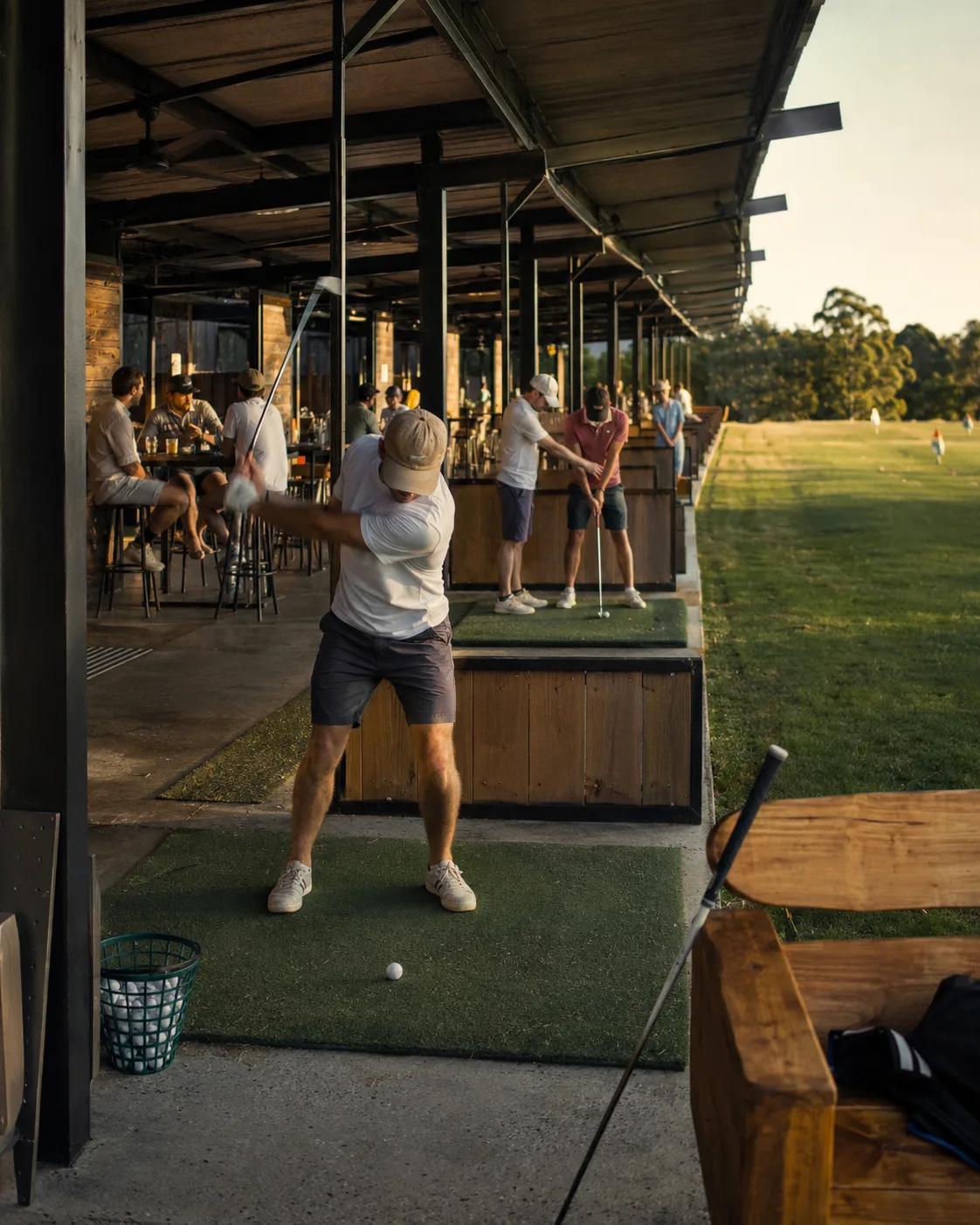 A golfer at the top of the backswing in an open-air range bay, with a bar patio and more bays behind